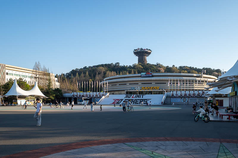 Korea-Changwon-Mall - The bike riding continues outside. Note that is the strange tree of life thing on top of the hill behind the basketball stadium.