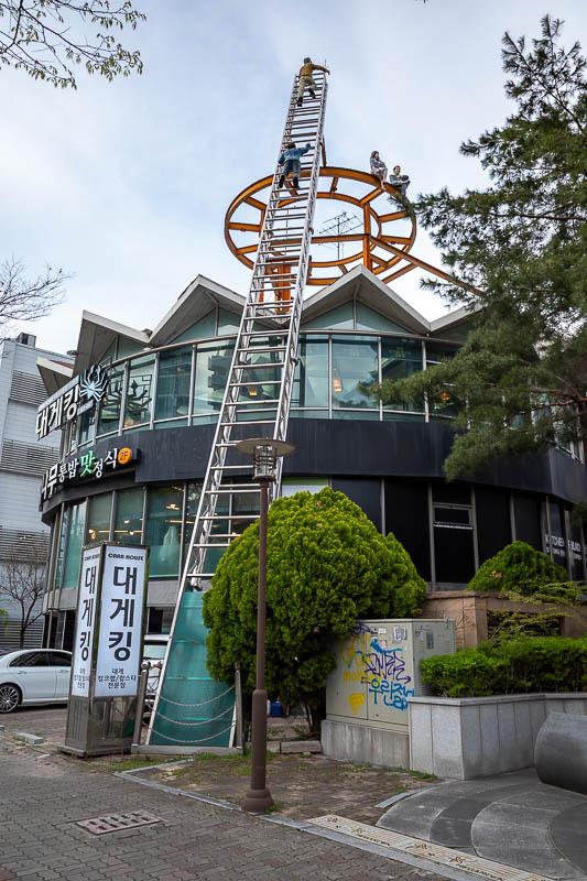 Korea-Daejeon-Shinsegae - This ladder goes to the roof of the crab house, for no reason at all. There are oversized human figurines playing on the ladder and the roof. The plas